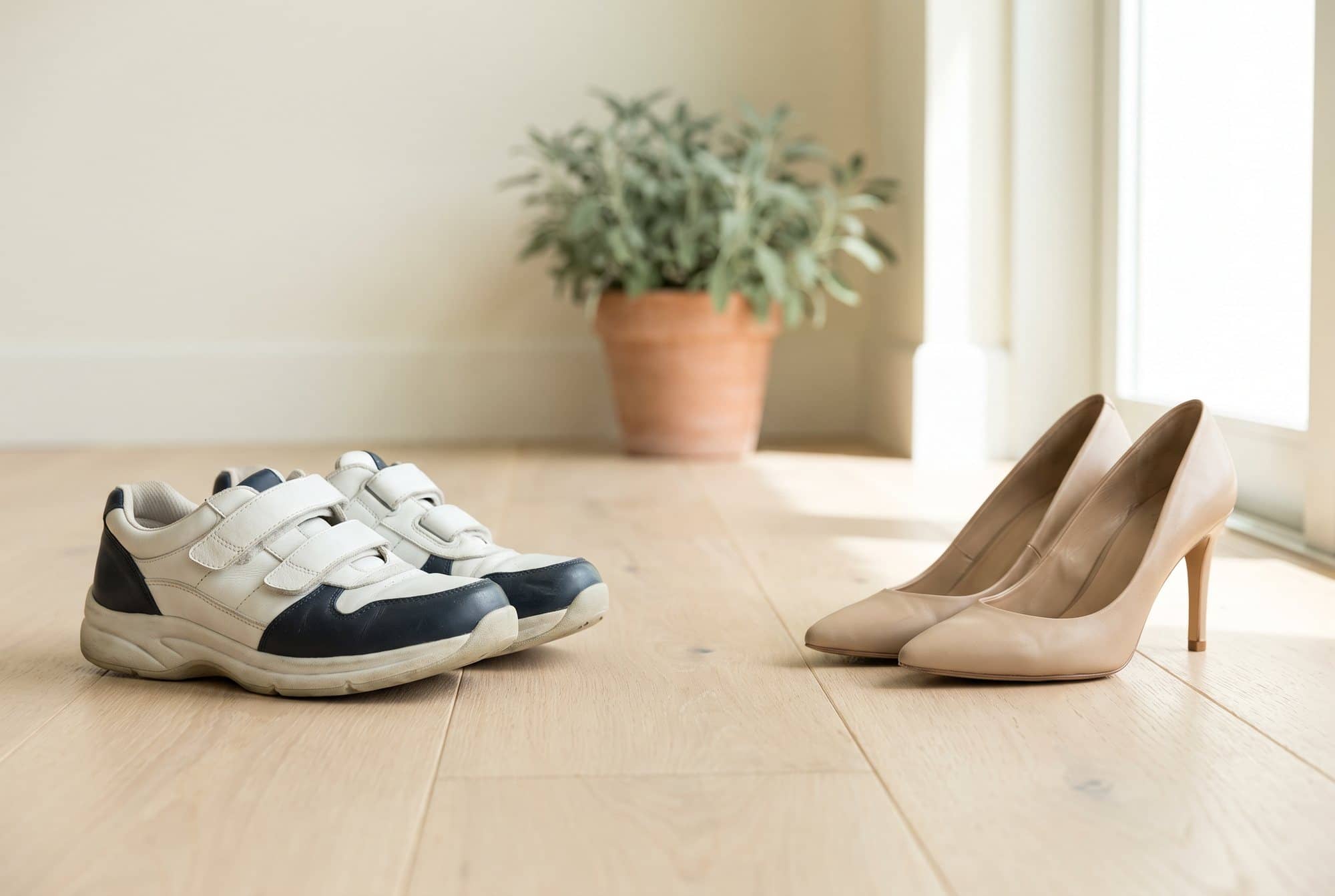 Supportive white and navy walking shoes with a wide toe box beside a pair of narrow, pointed beige high heels on a light oak floor