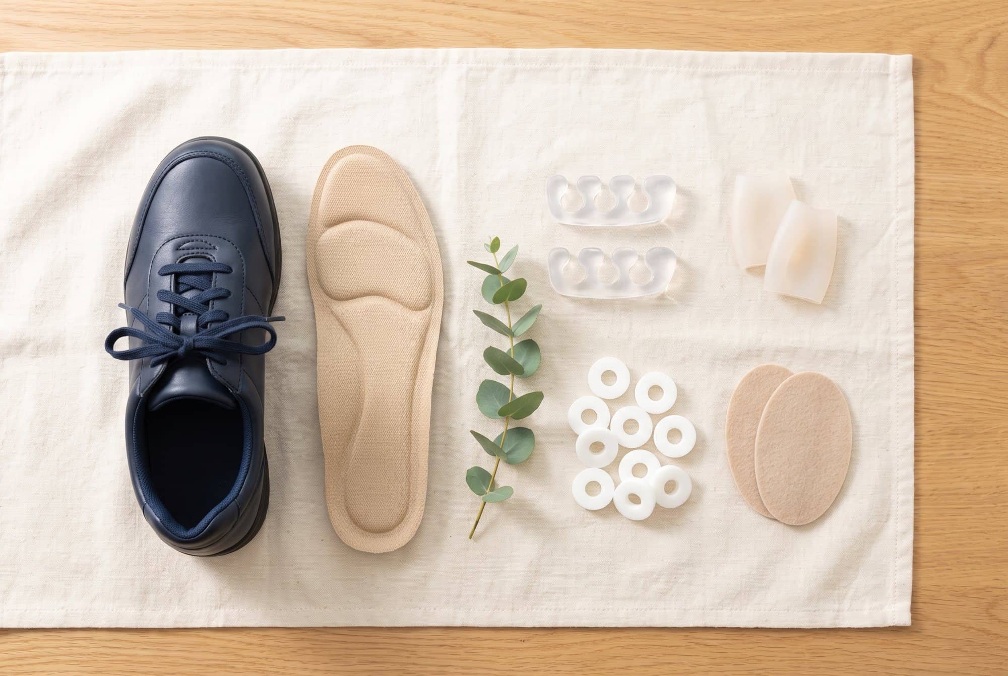 Flat lay of a navy walking shoe, a cushioned insole, clear gel toe separators, silicone toe sleeves, white foam ring pads and oval felt pads on ivory linen