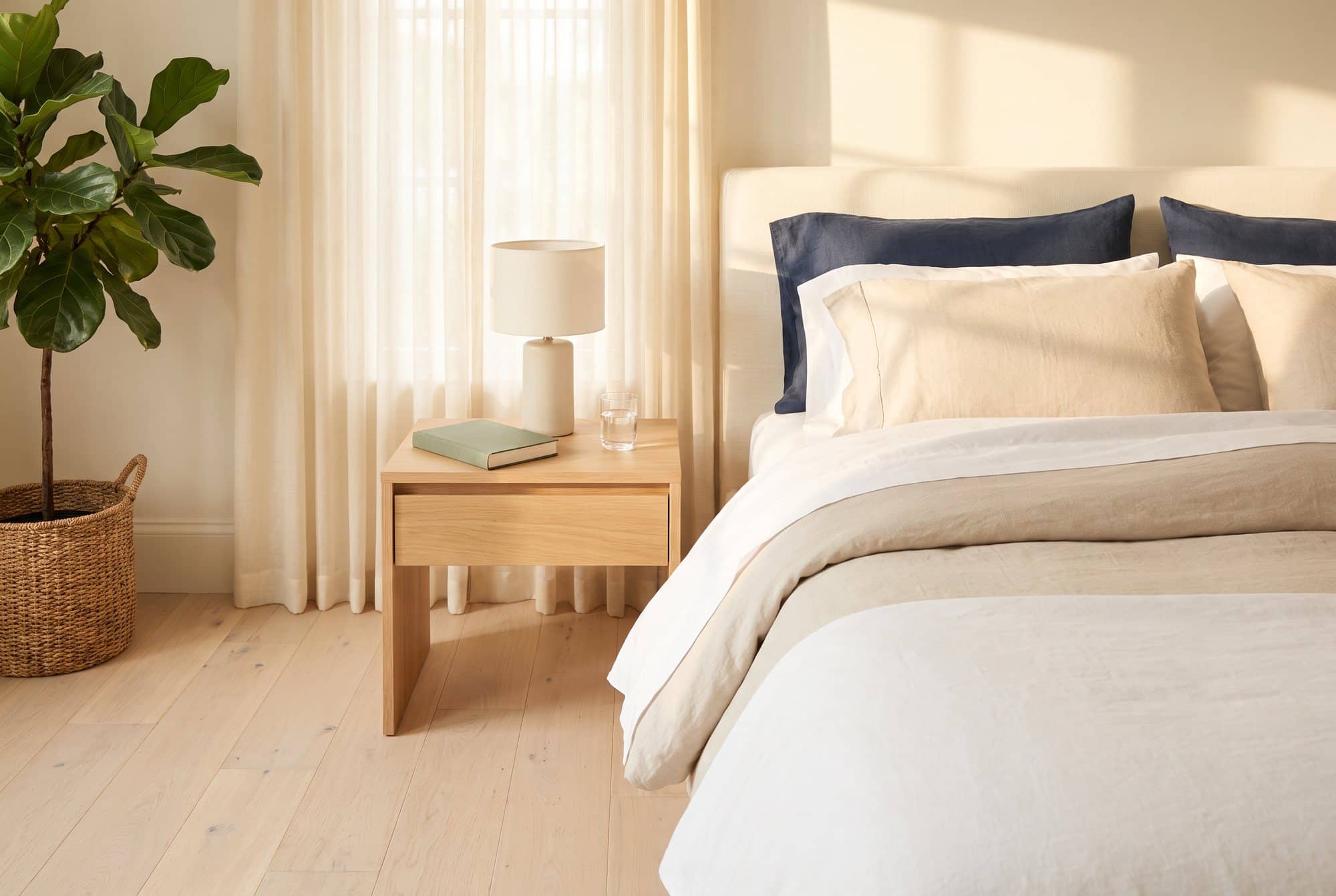 A calm bedroom at sunrise with a neatly made bed, sheer curtains, an oak nightstand with a lamp, a book and a glass of water