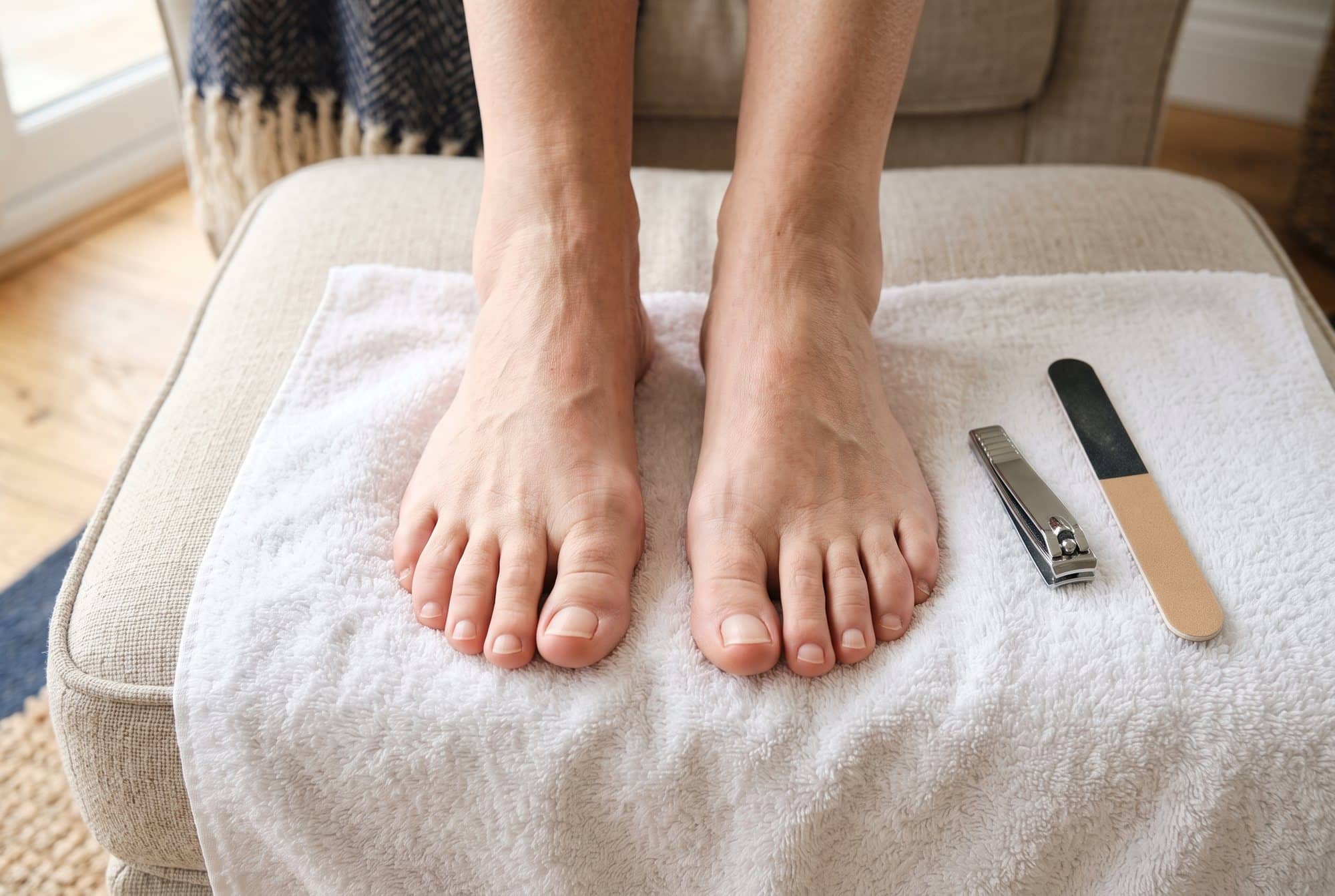Older adult's bare feet resting on a white towel on a footstool, toenails neatly trimmed straight across, with a toenail clipper and emery board beside them