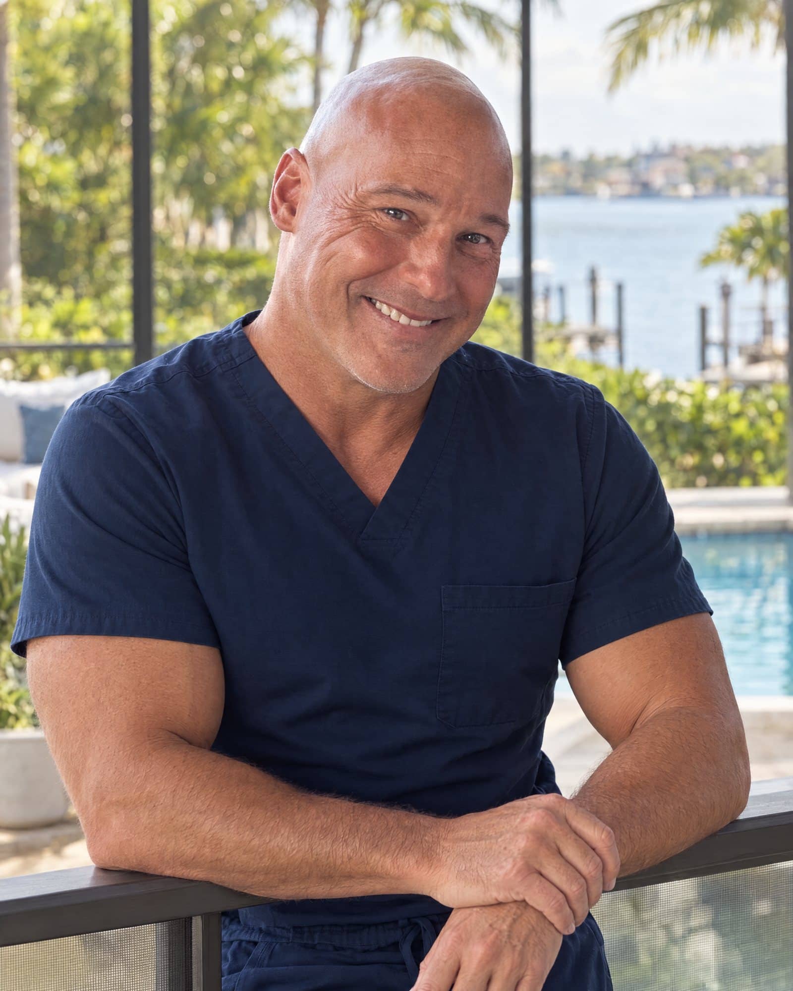 Dr. Robert Losier in navy scrubs, smiling as he leans on a lanai railing with the Naples waterfront behind him