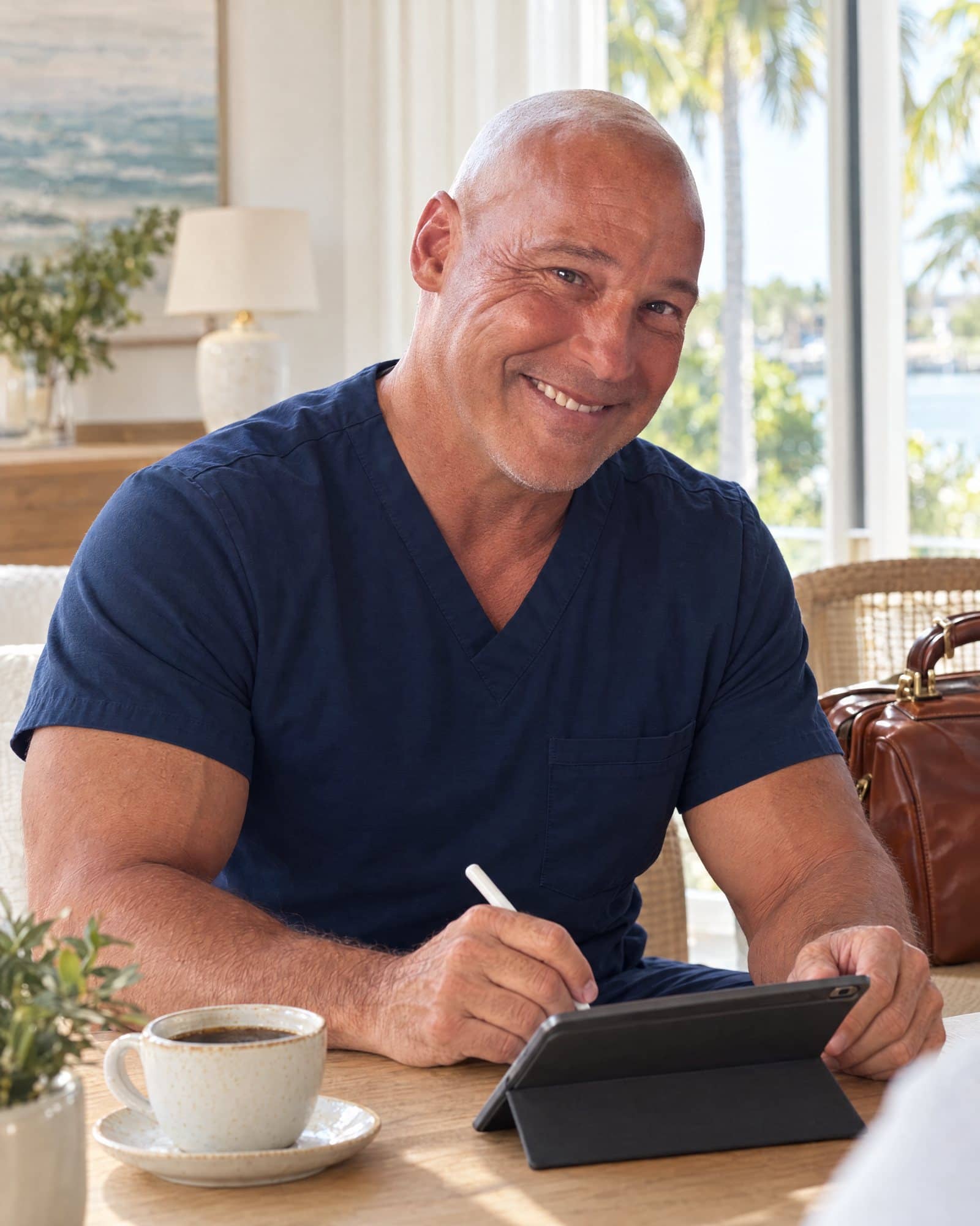 Dr. Losier smiling at a dining table as he writes visit notes on a tablet, a cup of coffee beside him